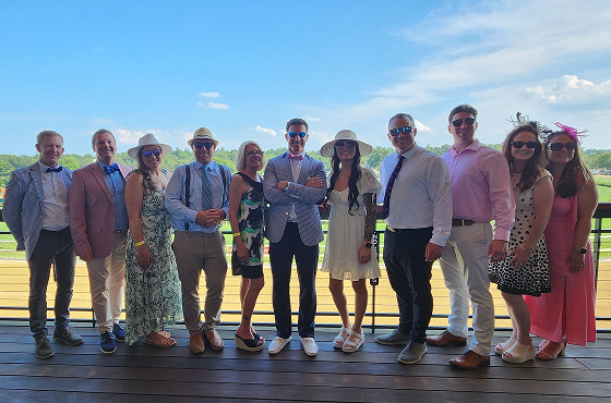 A group of adults in summer outfits posing for a photo on a wooden deck outdoors with a clear blue sky and sunshine.