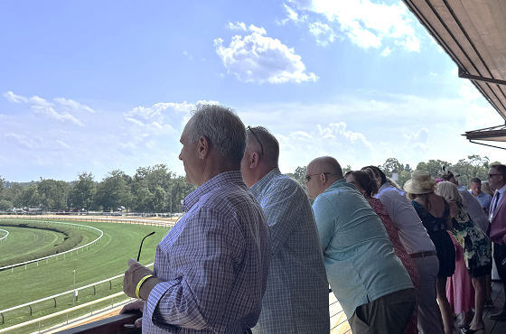 Several people lean on a railing and look out over a green racecourse on a sunny day, suggesting a spectator viewing area at an event.